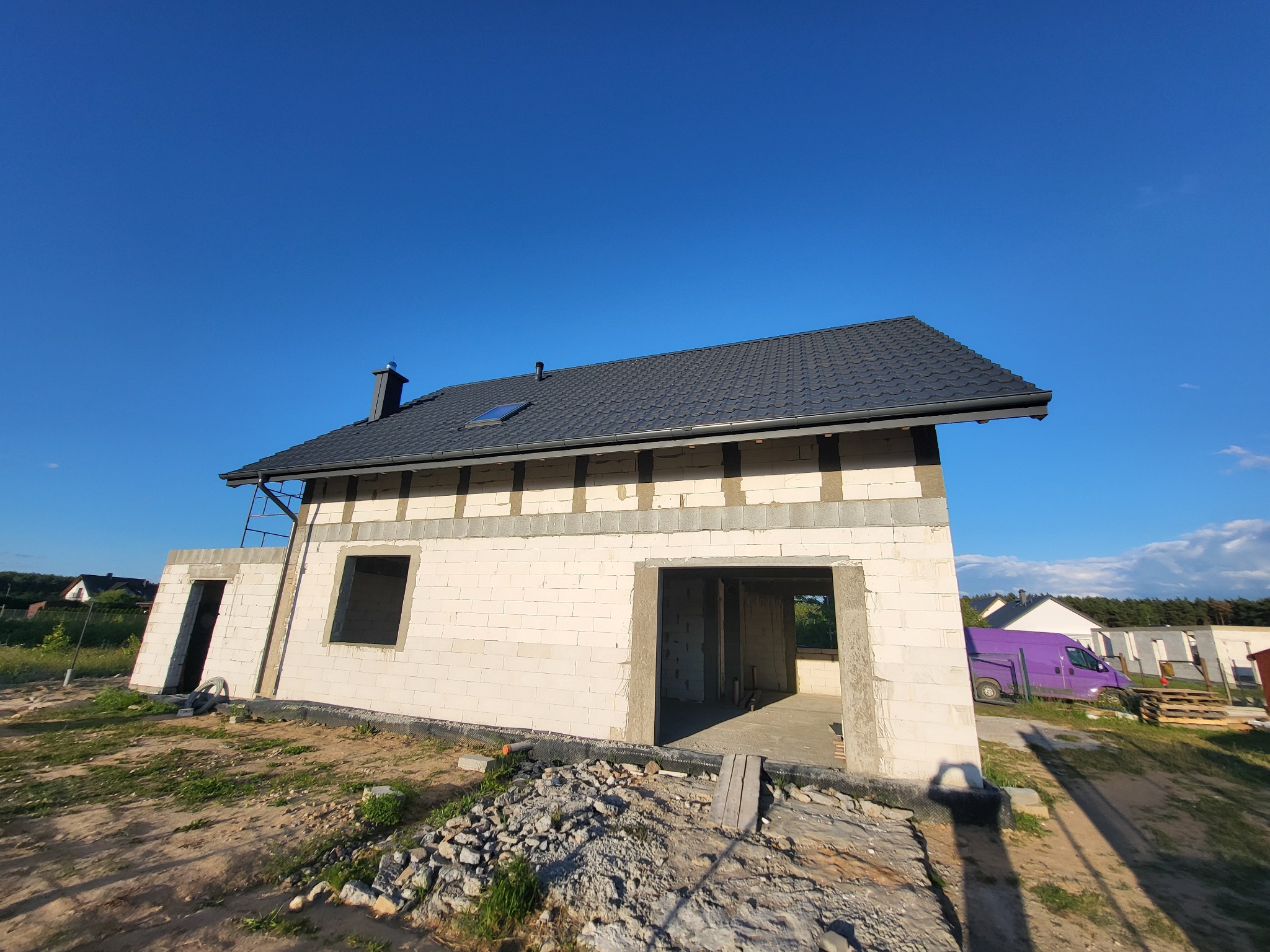 Niedokończony dom murowany z czarną dachówką i widocznym kominem, białe ściany z betonu komórkowego, otoczenie placu budowy z fioletowym busem w tle.
