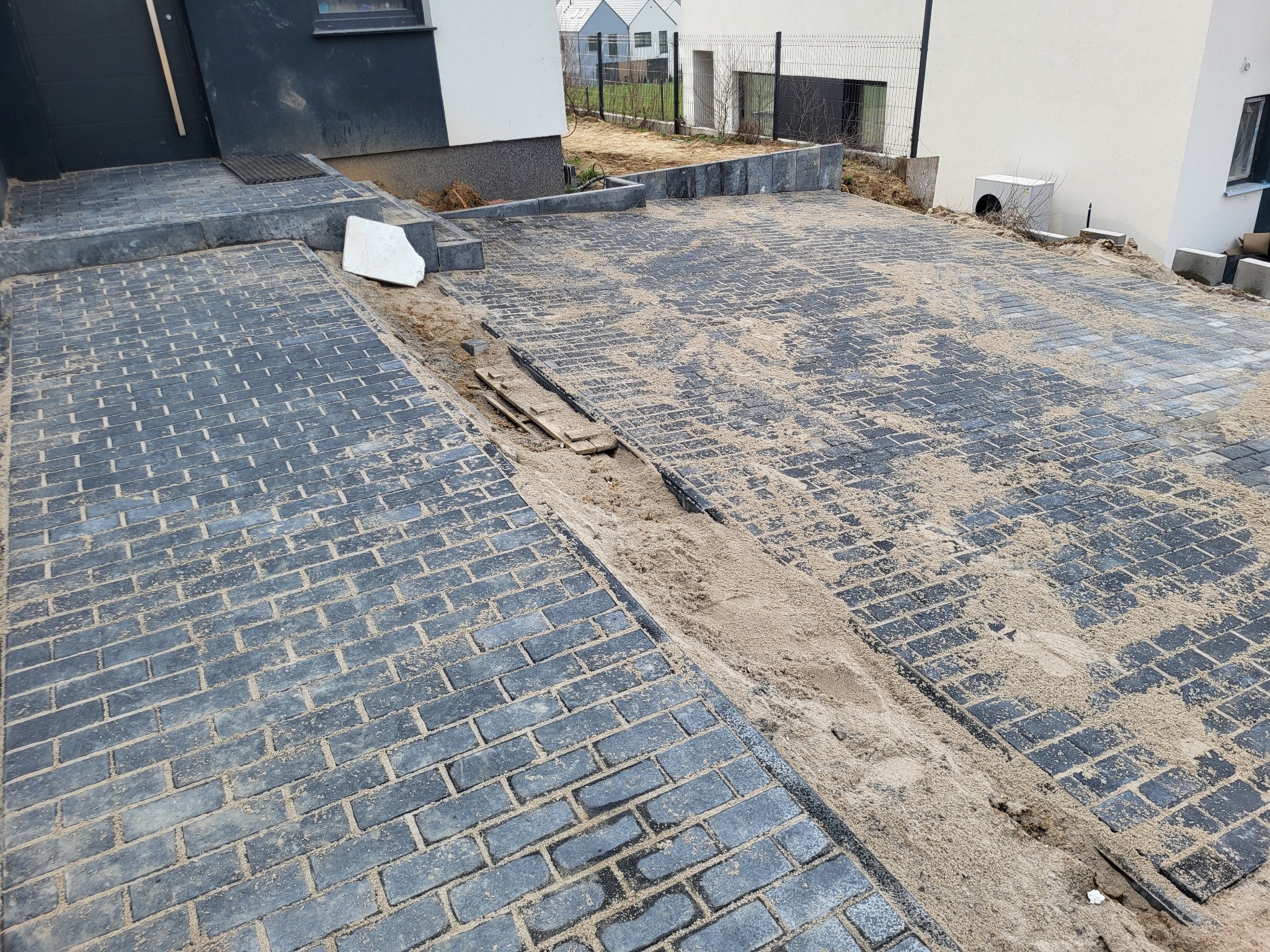 Ułożona kostka brukowa w kolorze grafitowym, zasypana piaskiem, w trakcie prac wykończeniowych na podjeździe przed domem. Widoczne niedociągnięcia i brakujące elementy.