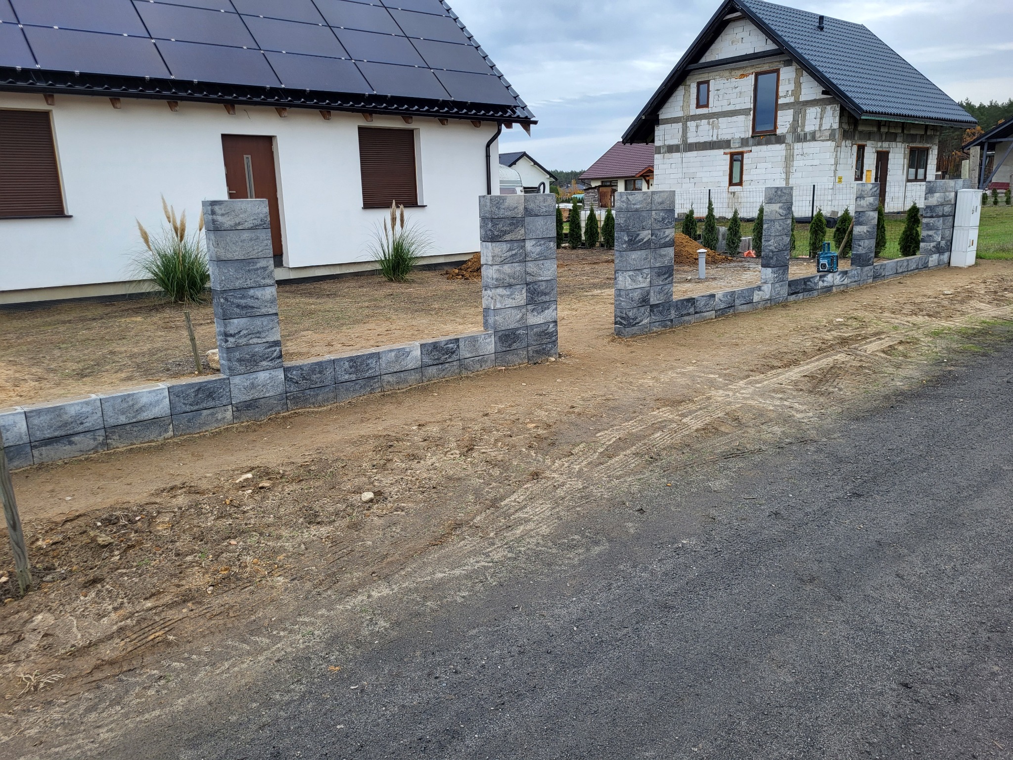 Wznoszenie ogrodzenia z szarej kostki modułowej na posesji z dwoma domami, jeden z panelami słonecznymi na dachu, drugi w trakcie budowy z białego pustaka.