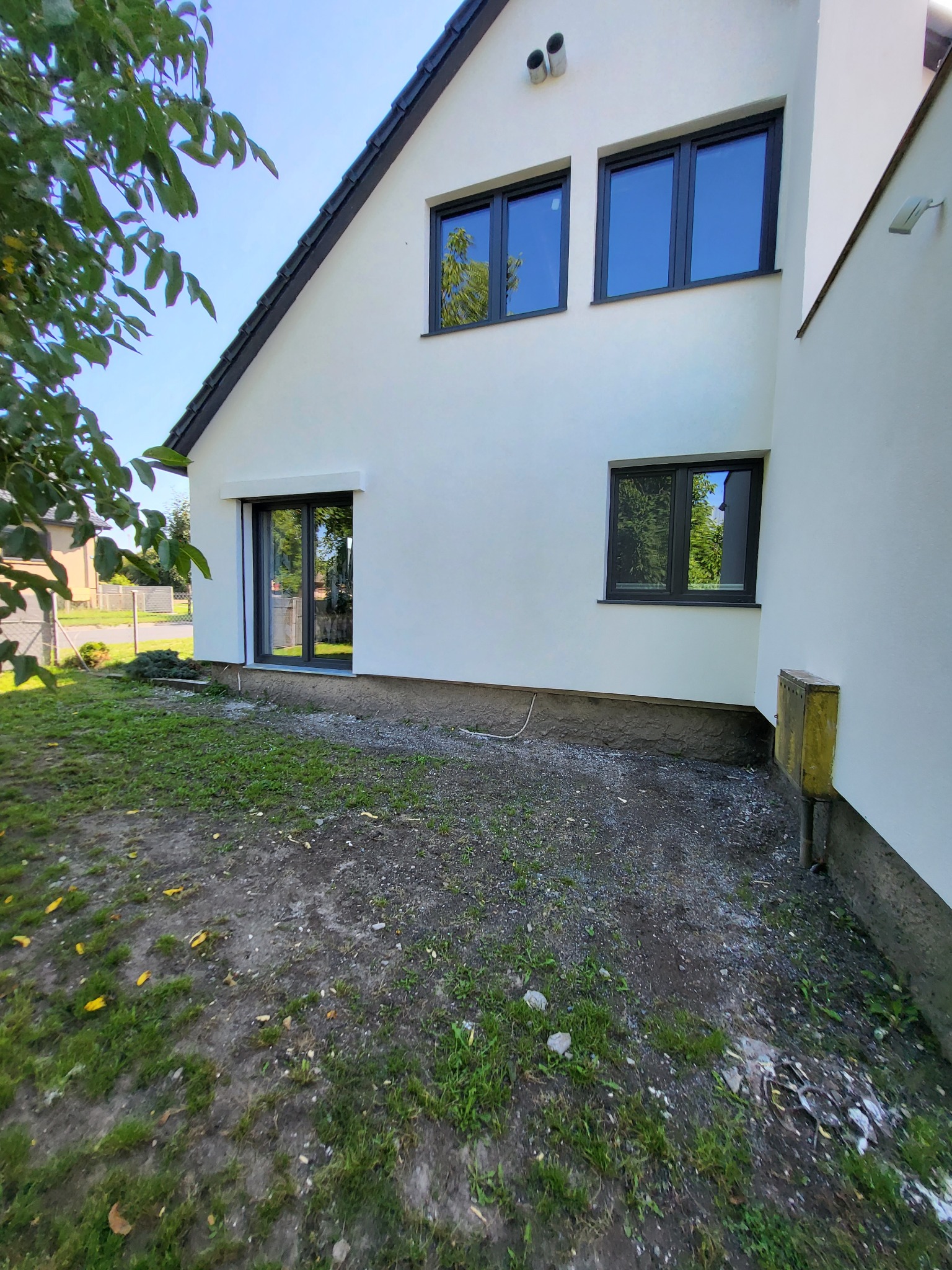 Świeżo otynkowany, biały dom jednorodzinny z czarną stolarką okienną i drzwiami tarasowymi, widok z boku na trawnik z resztkami gruzu.