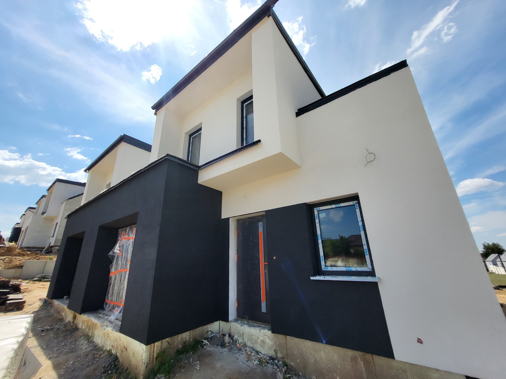 Nowoczesny dom w zabudowie szeregowej w Gębicach z czarno-białą elewacją, okna z niebieskimi ramami, drzwi zabezpieczone folią, widoczne niezagospodarowane otoczenie budynku.