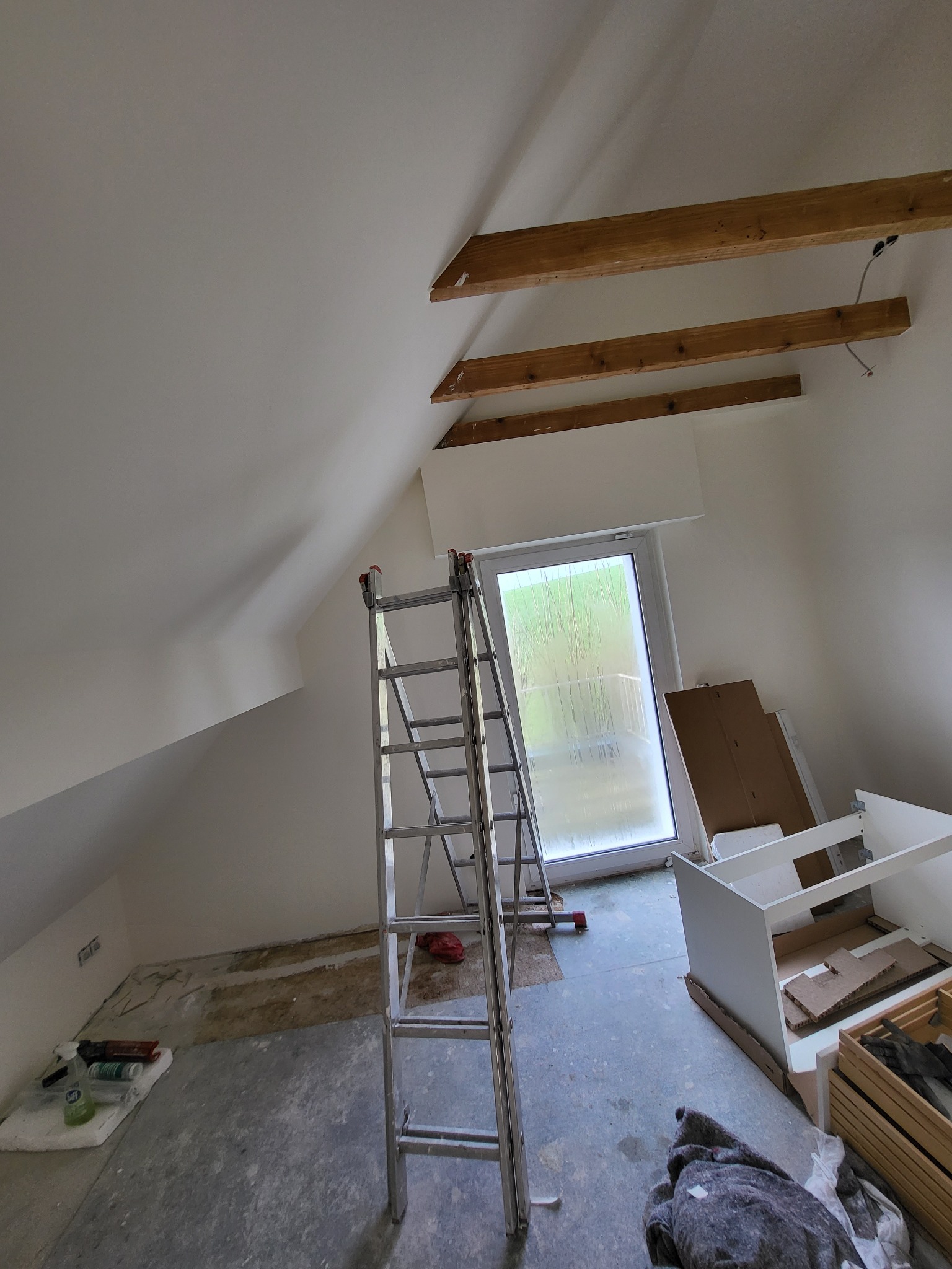 Poddasze w trakcie malowania: białe ściany, drewniane belki sufitowe, drabina aluminiowa, okno balkonowe, przygotowane narzędzia i materiały malarskie na podłodze.
