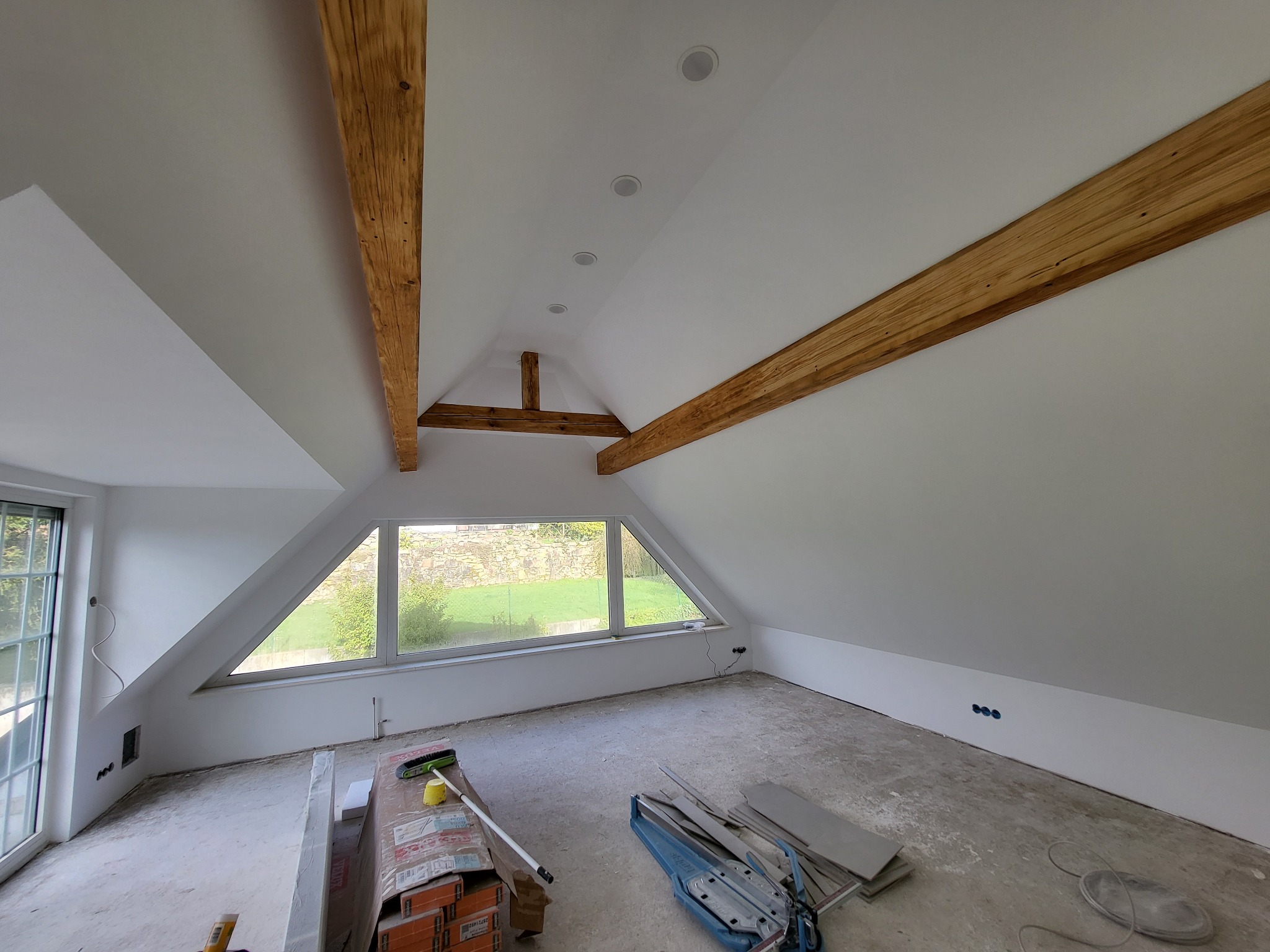 Poddasze w trakcie wykańczania z widocznymi drewnianymi belkami konstrukcyjnymi, oknem szczytowym i rozłożonymi narzędziami oraz materiałami budowlanymi na podłodze.