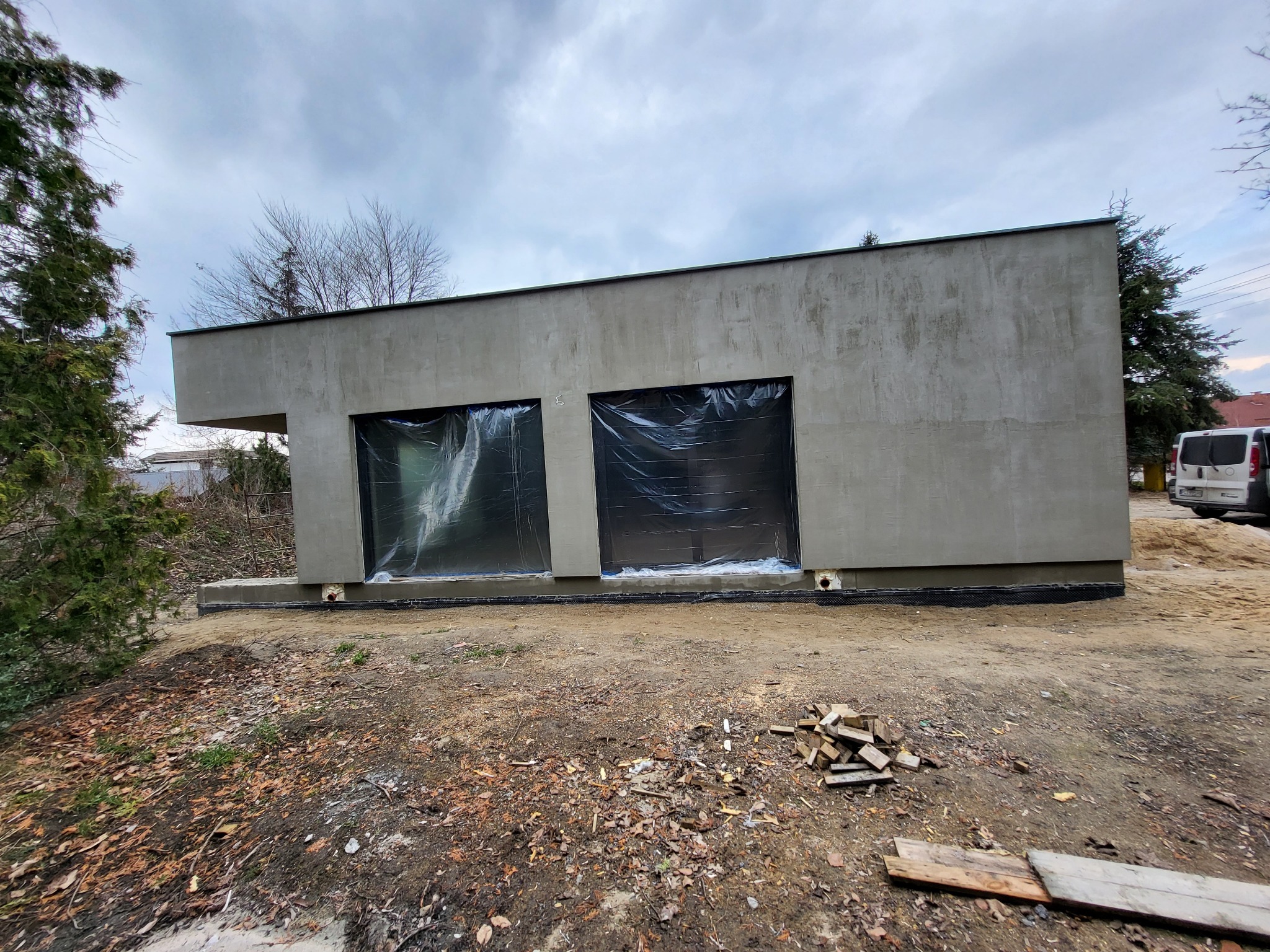 Surowy, betonowy budynek z dwoma oknami zabezpieczonymi folią, widoczne drewniane elementy na ziemi i fragment samochodu w tle, pochmurne niebo.