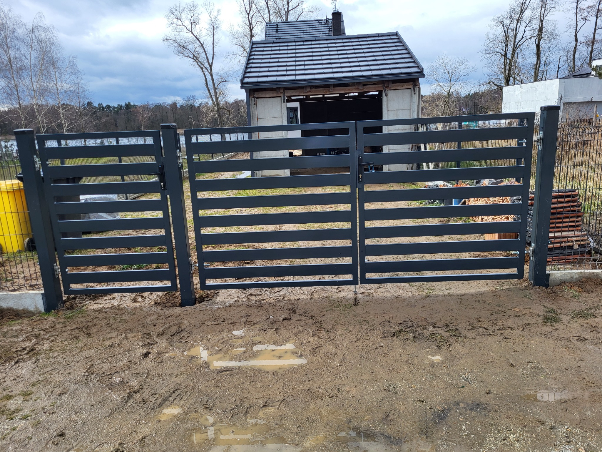 Nowoczesna, grafitowa brama wjazdowa z poziomymi panelami, widoczna z bliska, z błotnistym podłożem i budynkiem gospodarczym w tle.