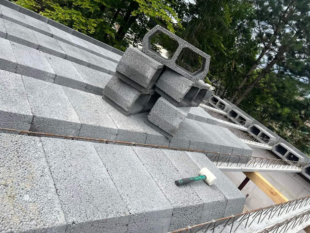 Wznoszenie ściany z szarych bloczków betonowych, widoczne zbrojenie i gumowy młotek, w tle zieleń drzew.