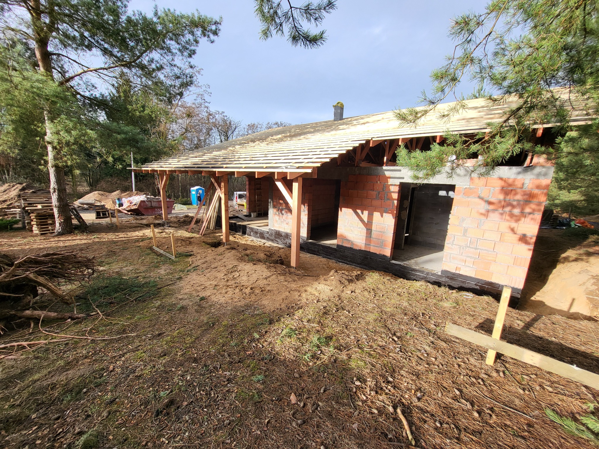 Budowa domu w lesie: surowe ściany z czerwonej cegły, drewniana konstrukcja dachu w trakcie montażu, widoczne rusztowania i materiały budowlane, otoczenie leśne z drzewami iglastymi.