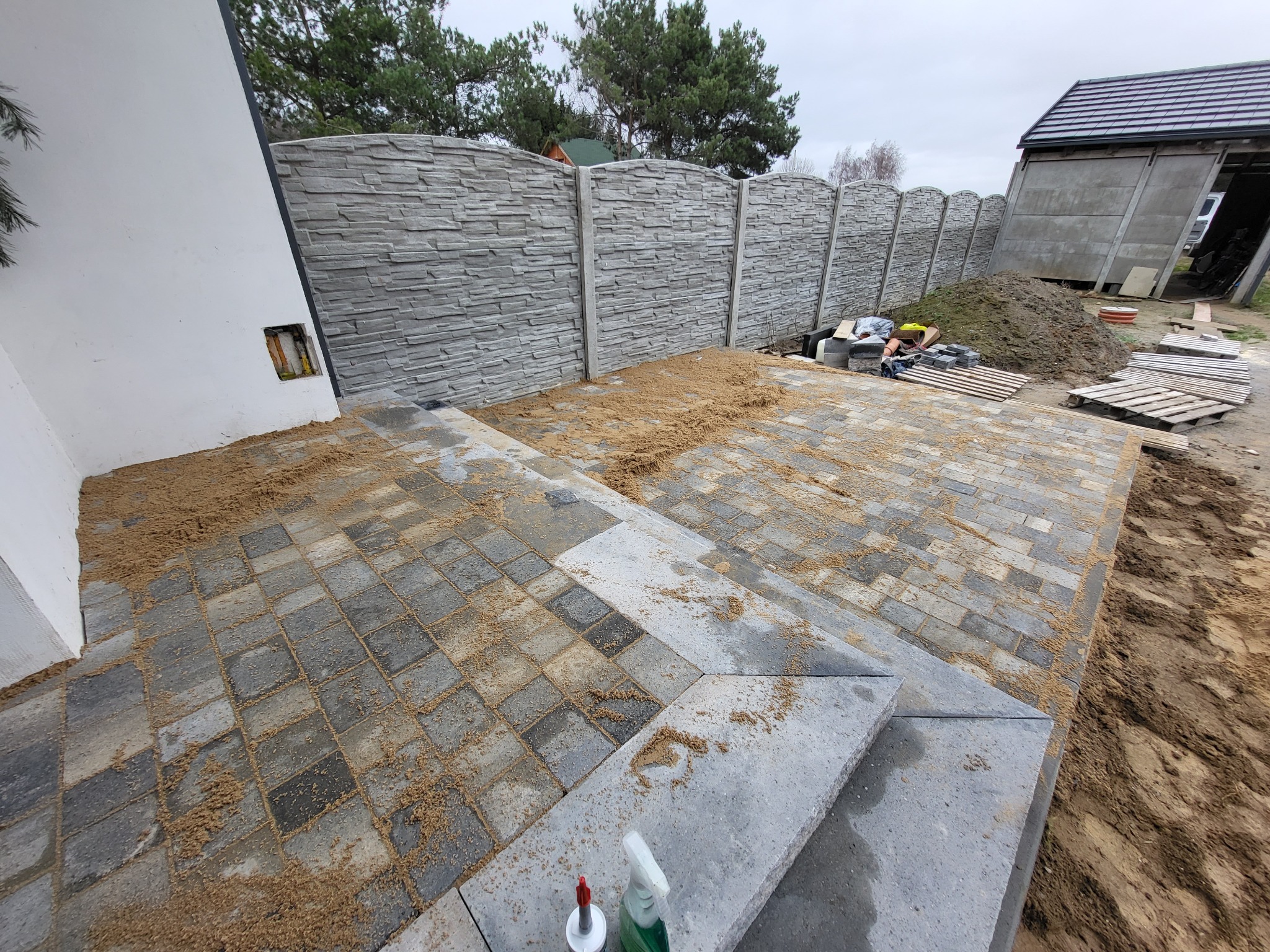 Montaż ogrodzenia z paneli imitujących kamień oraz układanie kostki brukowej na schodach i tarasie, widoczny piasek do fugowania, narzędzia i fragment domu gospodarczego w tle.