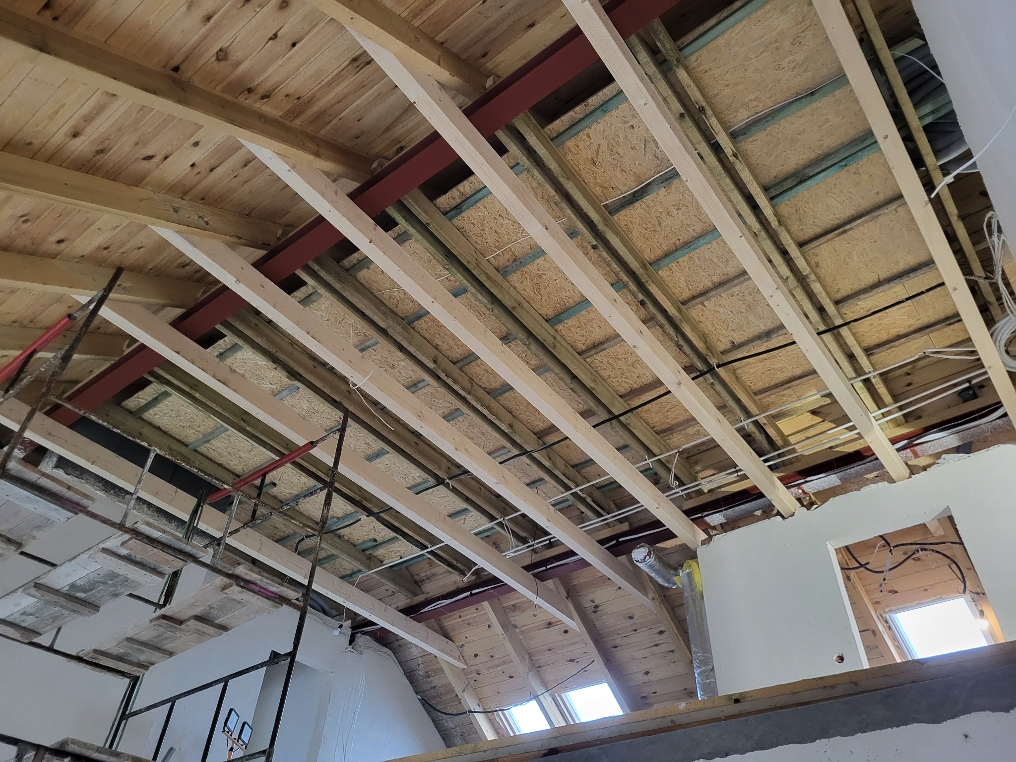 Wnętrze domu w trakcie rozbudowy: widok na drewniany strop z widocznymi belkami, metalowymi wzmocnieniami, instalacją elektryczną i fragmentem rusztowania.