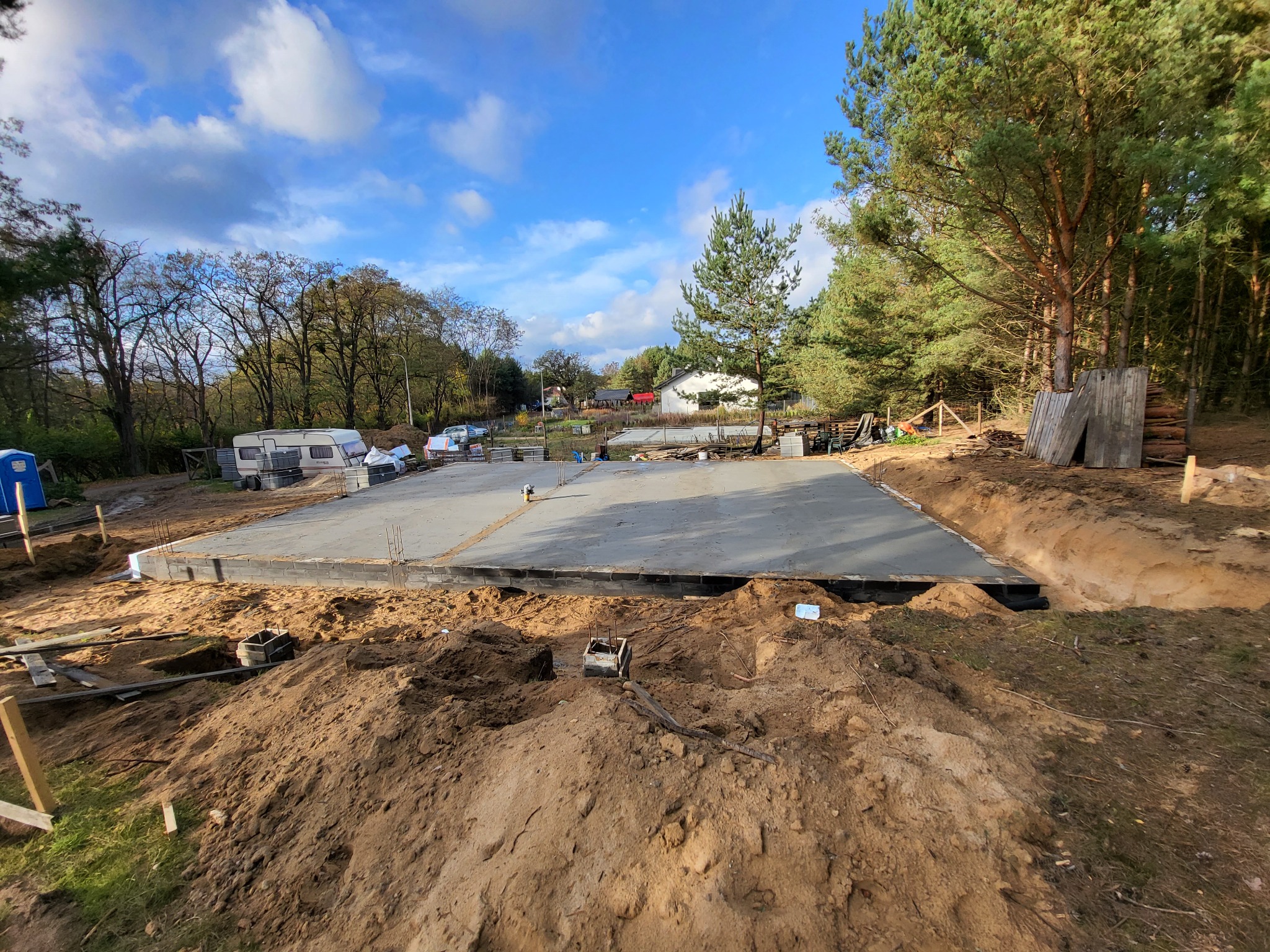 Wylana betonowa płyta fundamentowa pod budowę domu na działce z wykopami, widoczne szalunki i zbrojenia, w tle las i przyczepa kempingowa.
