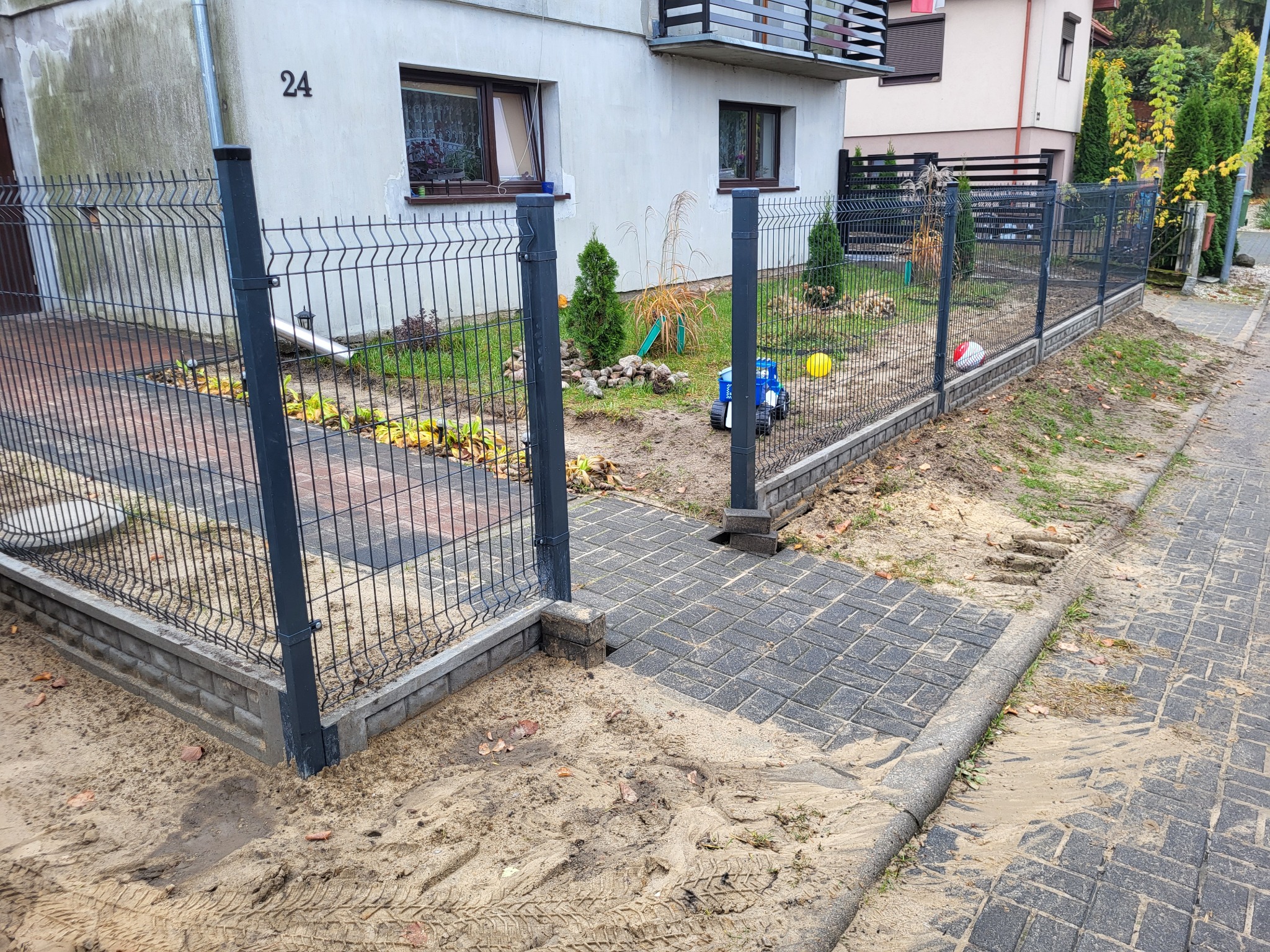 Nowo zainstalowane, metalowe ogrodzenie panelowe z podmurówką betonową, widoczne z zewnątrz posesji, z otwartą furtką i fragmentem chodnika z kostki brukowej, na tle dwukondygnacyjnego domu...