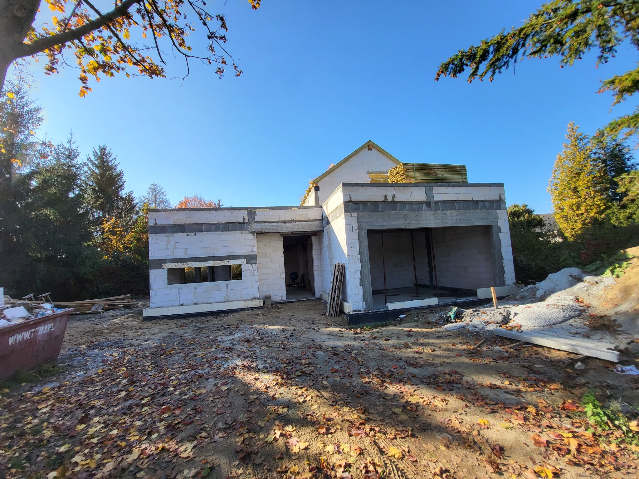 Surowy stan domu jednorodzinnego z garażem w trakcie budowy, ściany z betonu komórkowego, przygotowane drewno na dach, jesienne liście na ziemi.