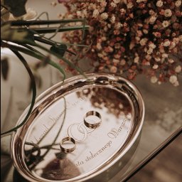 Czuła Fotografka - Dwa złote obrączki na srebrnej tacy z grawerem 'Urząd Stanu Cywilnego Miasta Stołecznego Warszawy', w tle bukiet z drobnymi kwiatami i różowa róża.