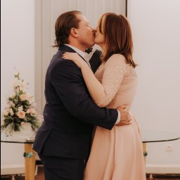 Czuła Fotografka - Para młoda w objęciach, pocałunek na tle jasnego wnętrza z kwiatami. Pan w granatowym garniturze, pani w pudrowej sukience z koronkowymi rękawami. Romantyczna fotografia ślubna.