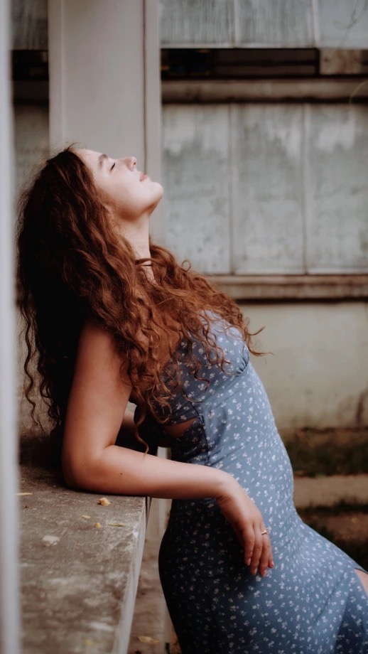 Portret kobiety z rudymi kręconymi włosami w błękitnej sukience w kwiaty, oparta o kamienny murek, z głową odchyloną do tyłu, w naturalnym oświetleniu.