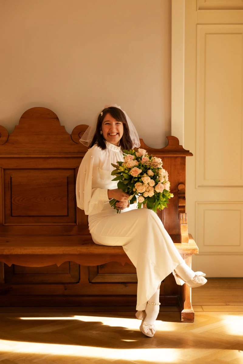 Uśmiechnięta panna młoda w białej sukni siedzi na drewnianej ławce, trzymając bukiet róż. Delikatny welon i jasne światło podkreślają radosny nastrój.