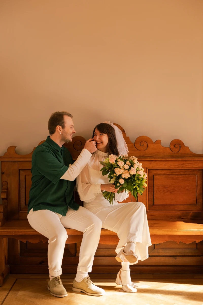 Uśmiechnięta para młoda siedzi na drewnianej ławce. Panna młoda w białym kombinezonie trzyma bukiet różowych kwiatów, pan młody w zielonej koszuli dotyka jej policzka.
