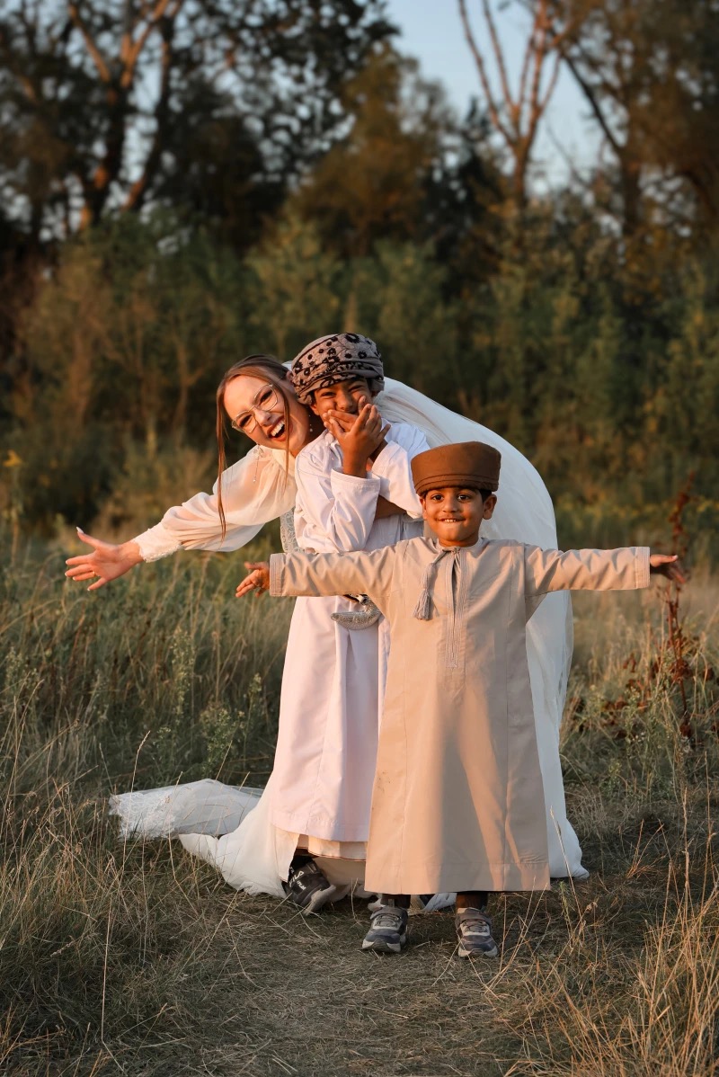 Radosna sesja rodzinna w plenerze: mama w białej sukience i dwóch synów w tradycyjnych strojach, pozują z rozłożonymi ramionami na tle zieleni.