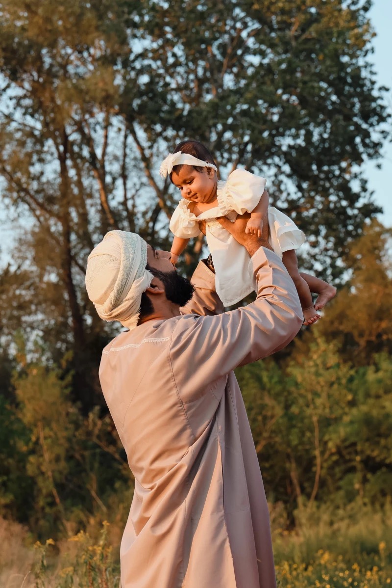 Uśmiechnięty tata w tradycyjnym stroju podnosi w górę małą córeczkę w białej sukience na tle zieleni. Ciepłe, naturalne światło podkreśla rodzinny moment.