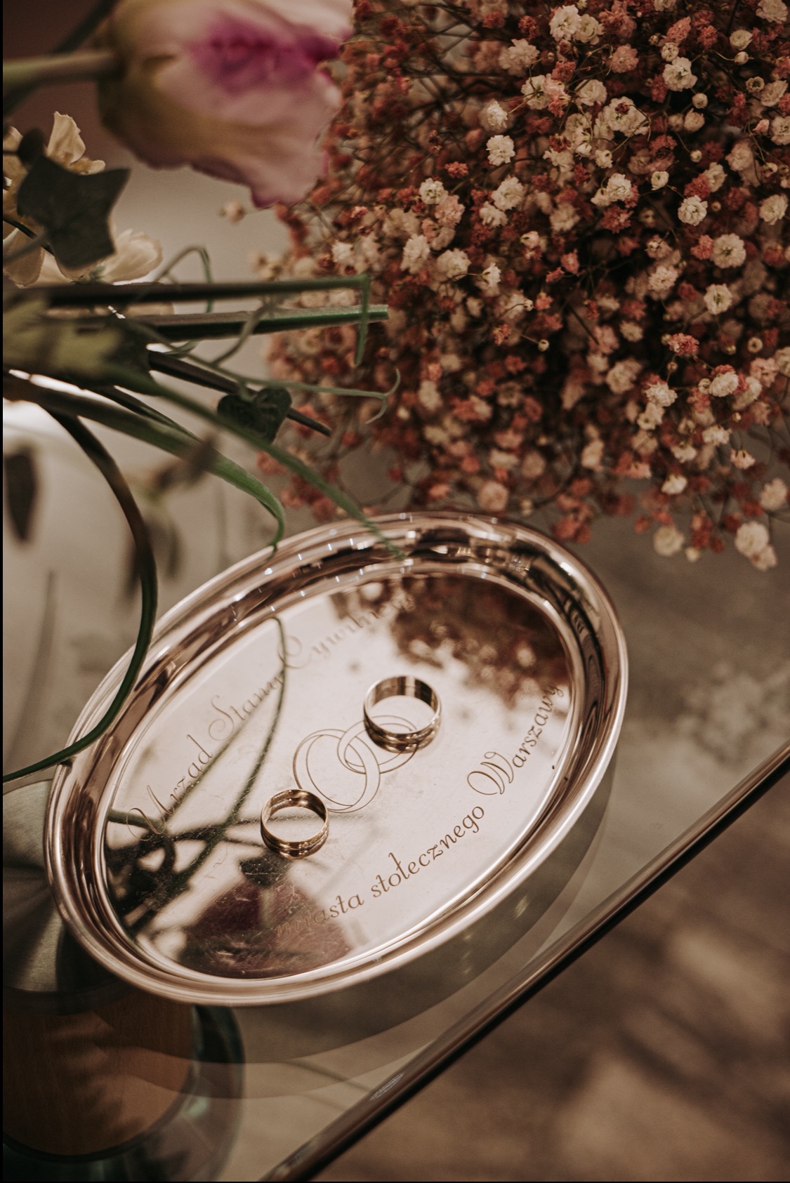 Dwa złote obrączki na srebrnej tacy z grawerem 'Urząd Stanu Cywilnego Miasta Stołecznego Warszawy', w tle bukiet z drobnymi kwiatami i różowa róża.