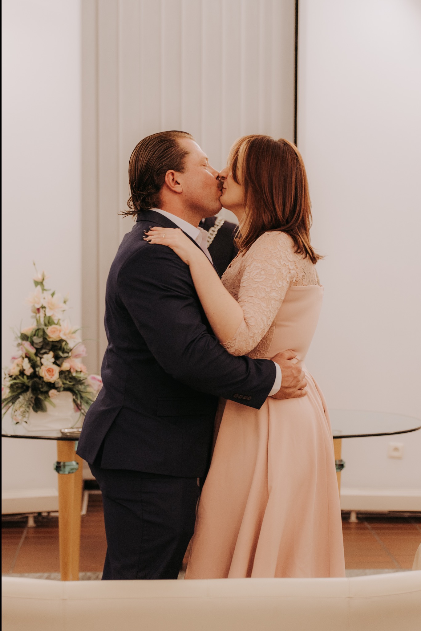 Para młoda w objęciach, pocałunek na tle jasnego wnętrza z kwiatami. Pan w granatowym garniturze, pani w pudrowej sukience z koronkowymi rękawami. Romantyczna fotografia ślubna.