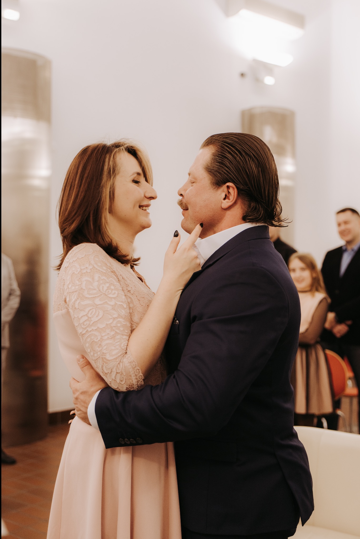 Uśmiechnięta para w objęciach podczas uroczystości, kobieta dotyka brody mężczyzny. W tle goście i jasne wnętrze.