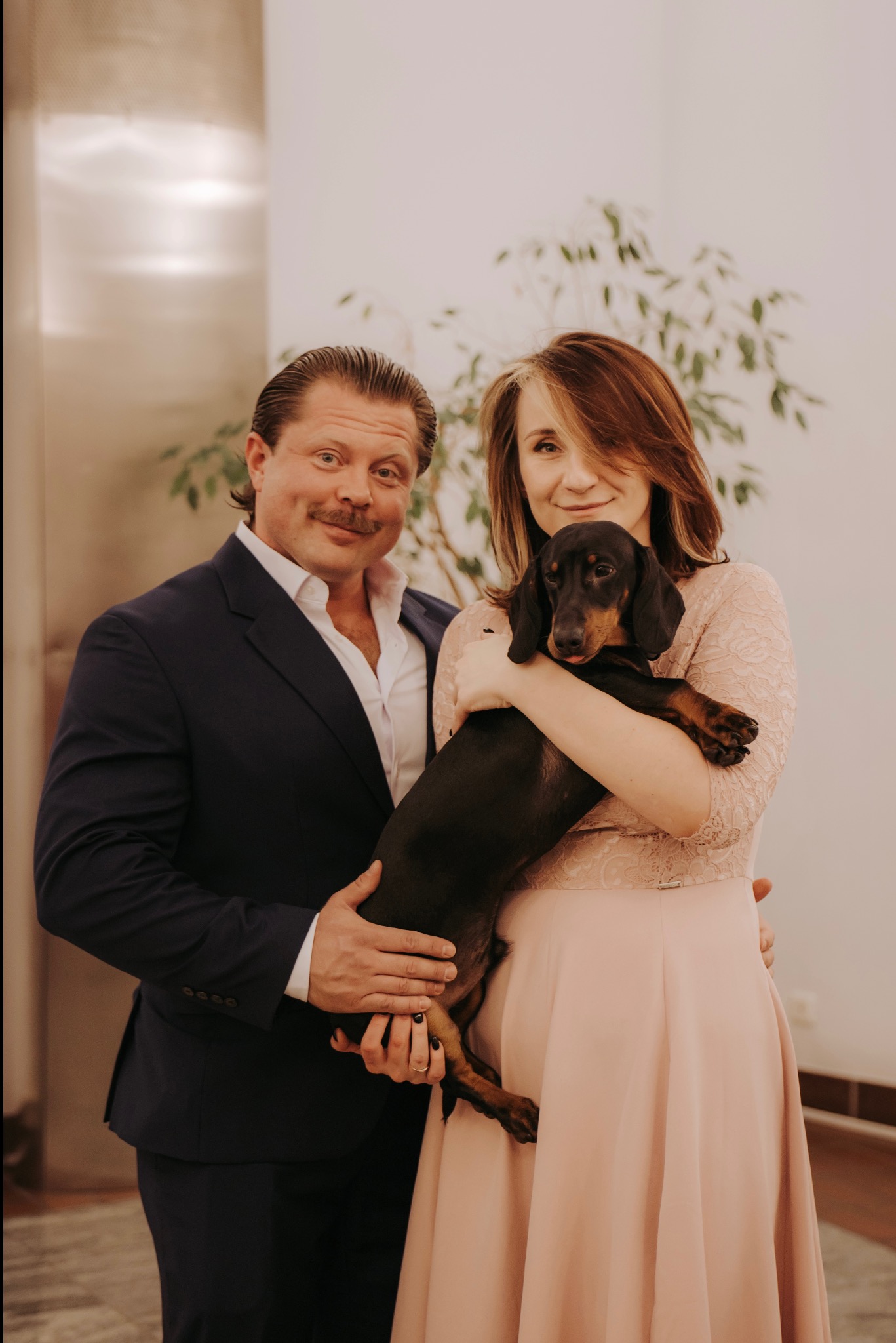 Uśmiechnięta para w eleganckich strojach pozuje z jamnikiem na tle jasnej ściany i rośliny doniczkowej. Mężczyzna w granatowym garniturze, kobieta w pudrowej sukience.