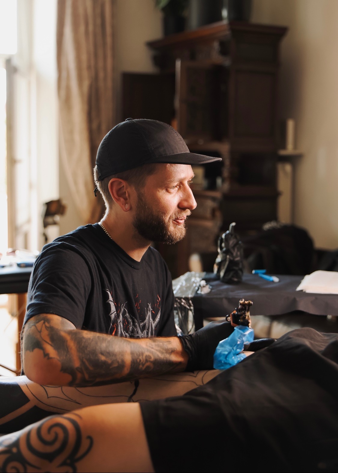 Tatuator w trakcie pracy nad tatuażem na nodze klienta w studiu w Warszawie. Widoczny wzór tatuażu, narzędzia i artysta w czarnej koszulce i czapce.