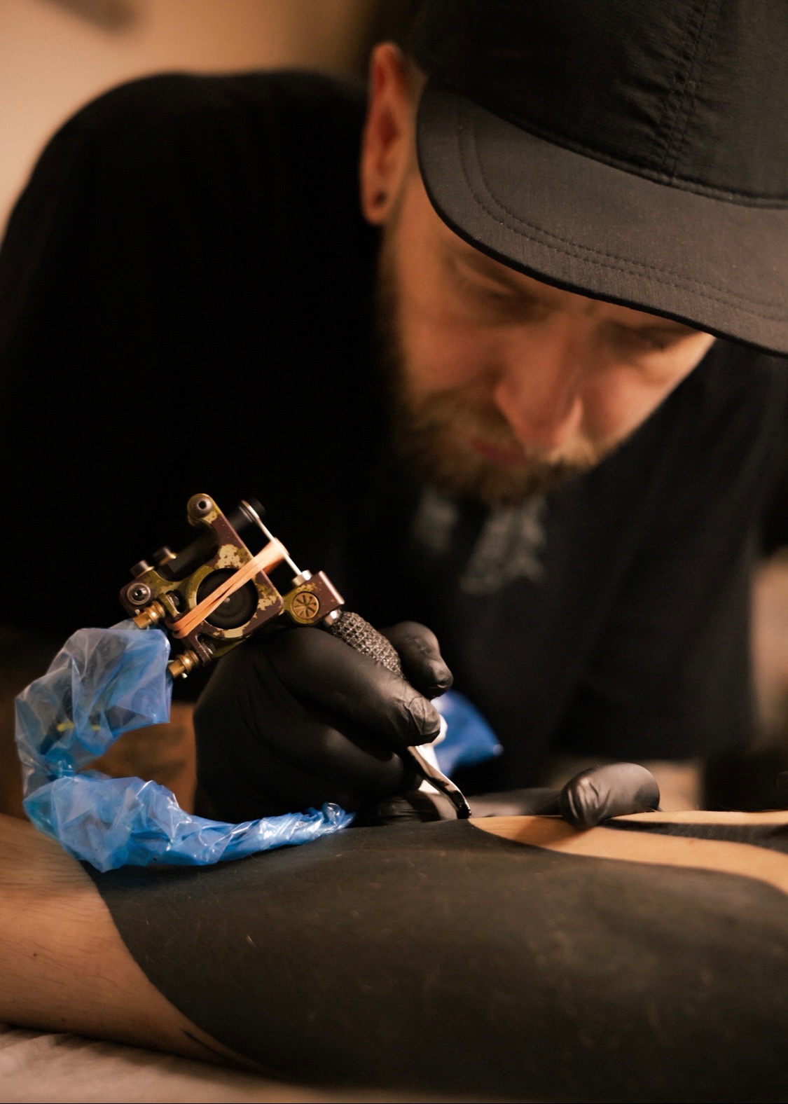 Tatuator w czarnych rękawiczkach wykonuje tatuaż blackwork na nodze klienta, używając maszynki z niebieskim zabezpieczeniem. Artysta w czapce skupiony na pracy.