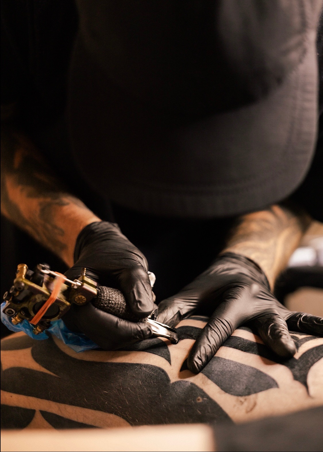 Tatuator w czarnych rękawiczkach wykonuje czarny tatuaż na skórze klienta, używając maszynki do tatuażu. Widoczne wytatuowane ręce artysty i fragment wzoru na ciele.