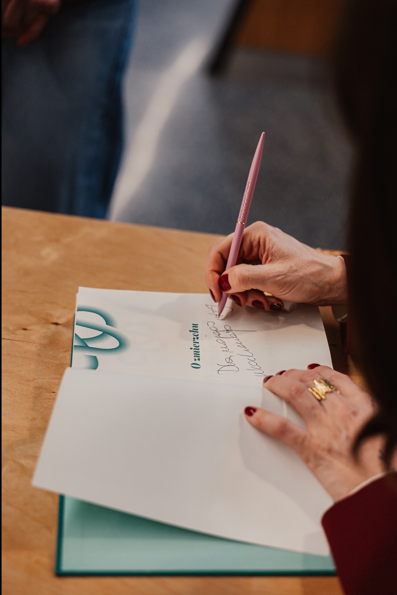 Osoba wpisuje dedykację do pamiątkowej księgi gości, używając różowego długopisu. Widoczny złoty pierścionek i czerwony lakier na paznokciach.