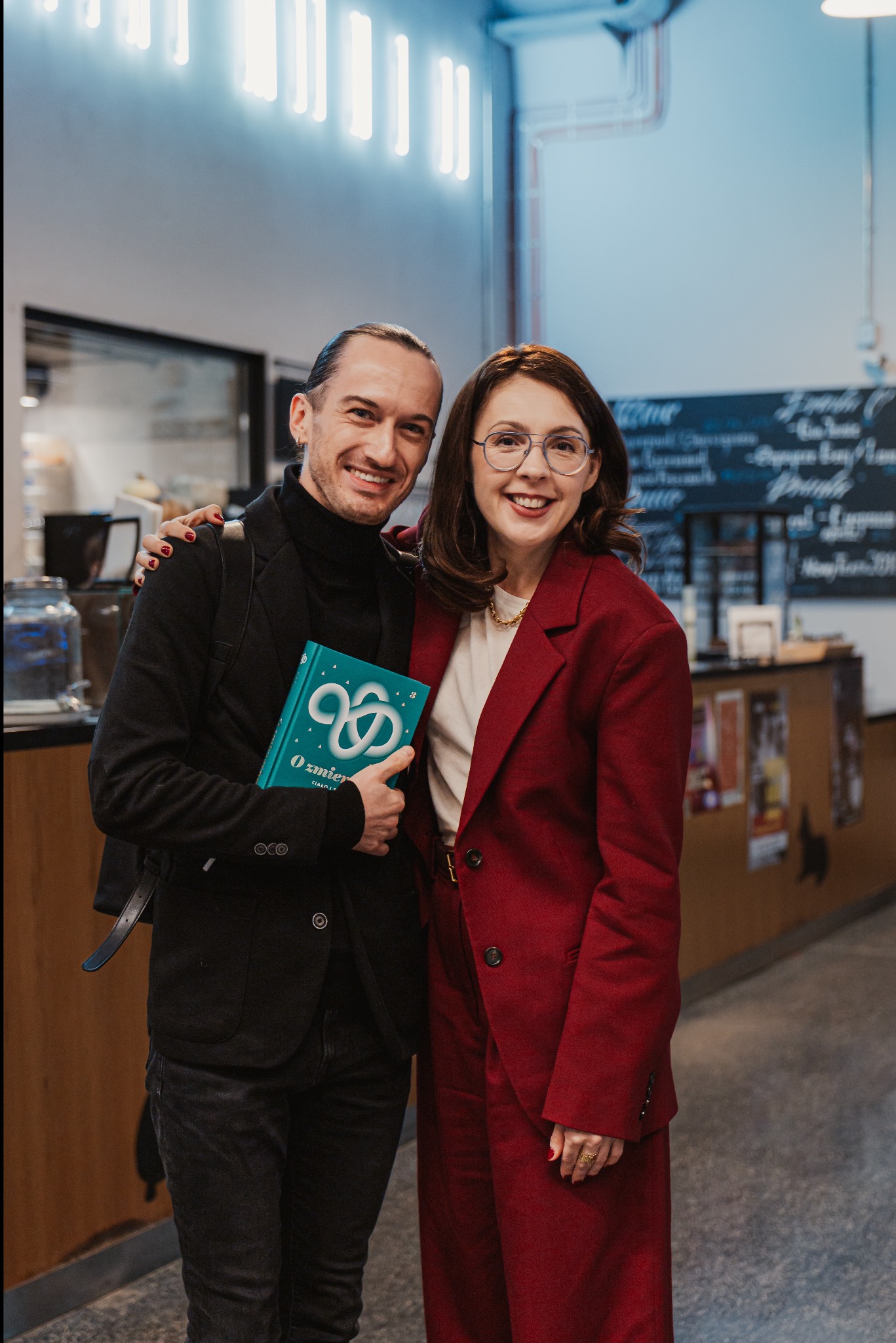Uśmiechnięta para w eleganckich strojach pozuje w kawiarni. Mężczyzna trzyma w ręku książkę z turkusową okładką. Kobieta w czerwonym garniturze i okularach.