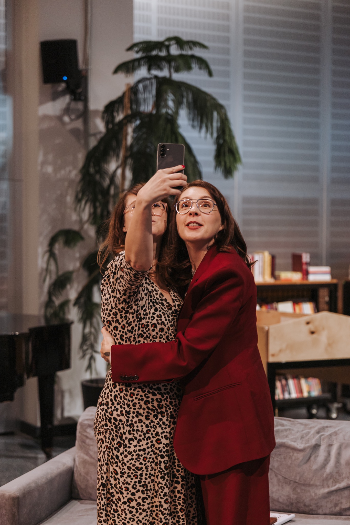 Dwie kobiety obejmują się, robiąc selfie telefonem. Jedna w sukience w panterkę, druga w czerwonym garniturze. W tle pianino, regał z książkami i roślina doniczkowa.