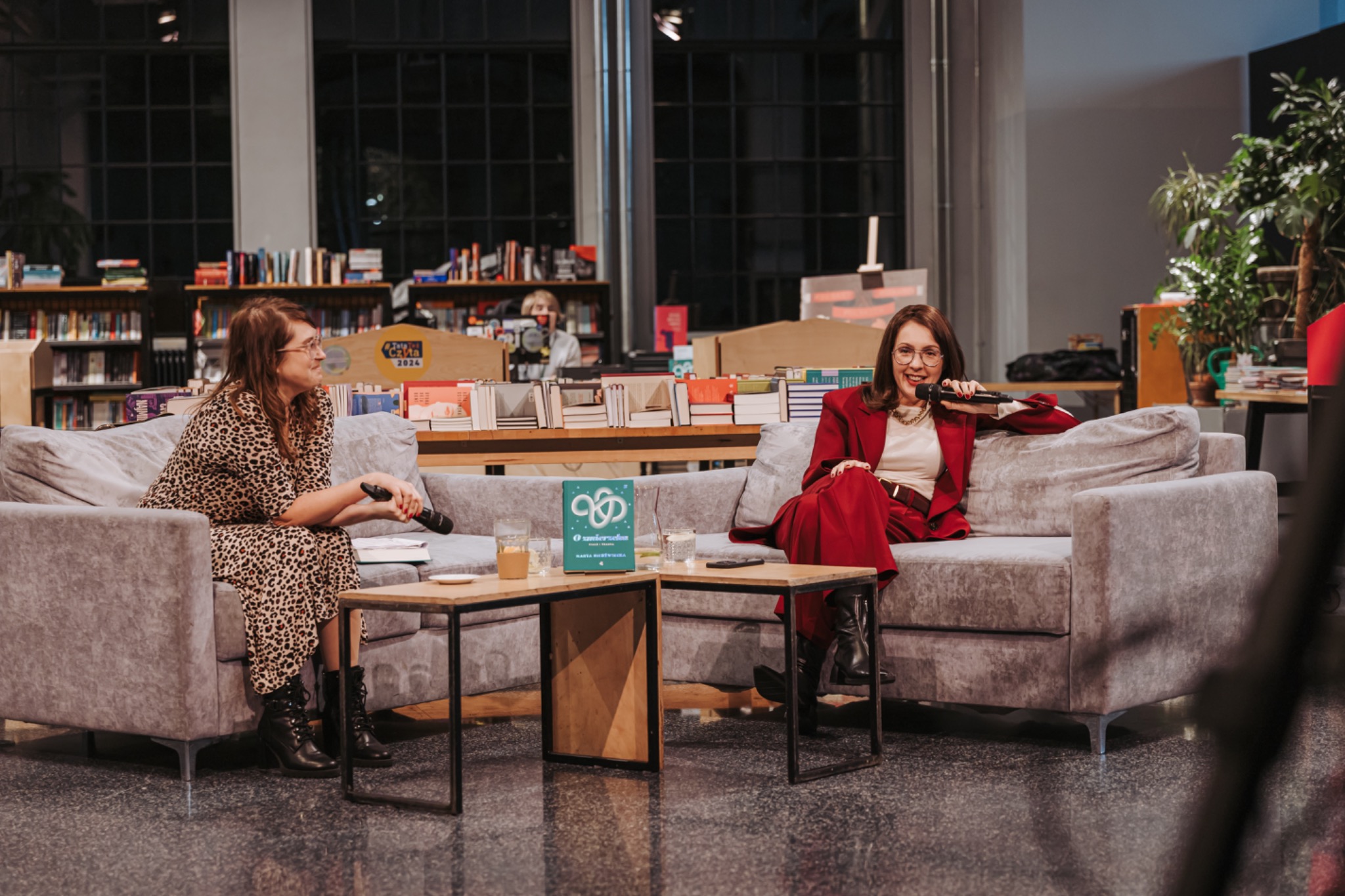 Dwie kobiety siedzą na kanapie podczas spotkania autorskiego w bibliotece. Jedna w czerwonym garniturze trzyma mikrofon. Na stoliku książka i napoje.