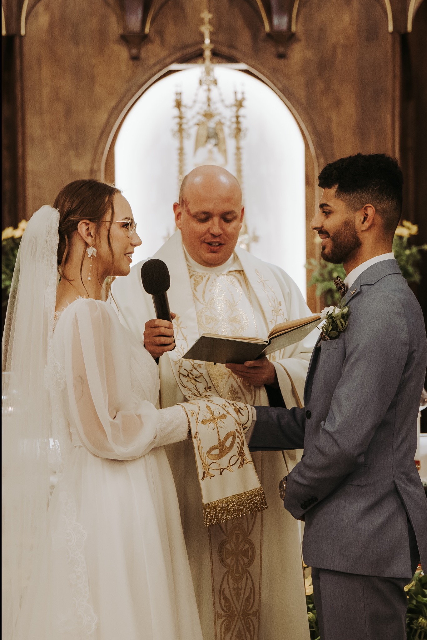 Uśmiechnięta para młoda podczas ceremonii ślubnej w kościele, trzymająca mikrofon i księgę, z widocznym księdzem w szatach liturgicznych.