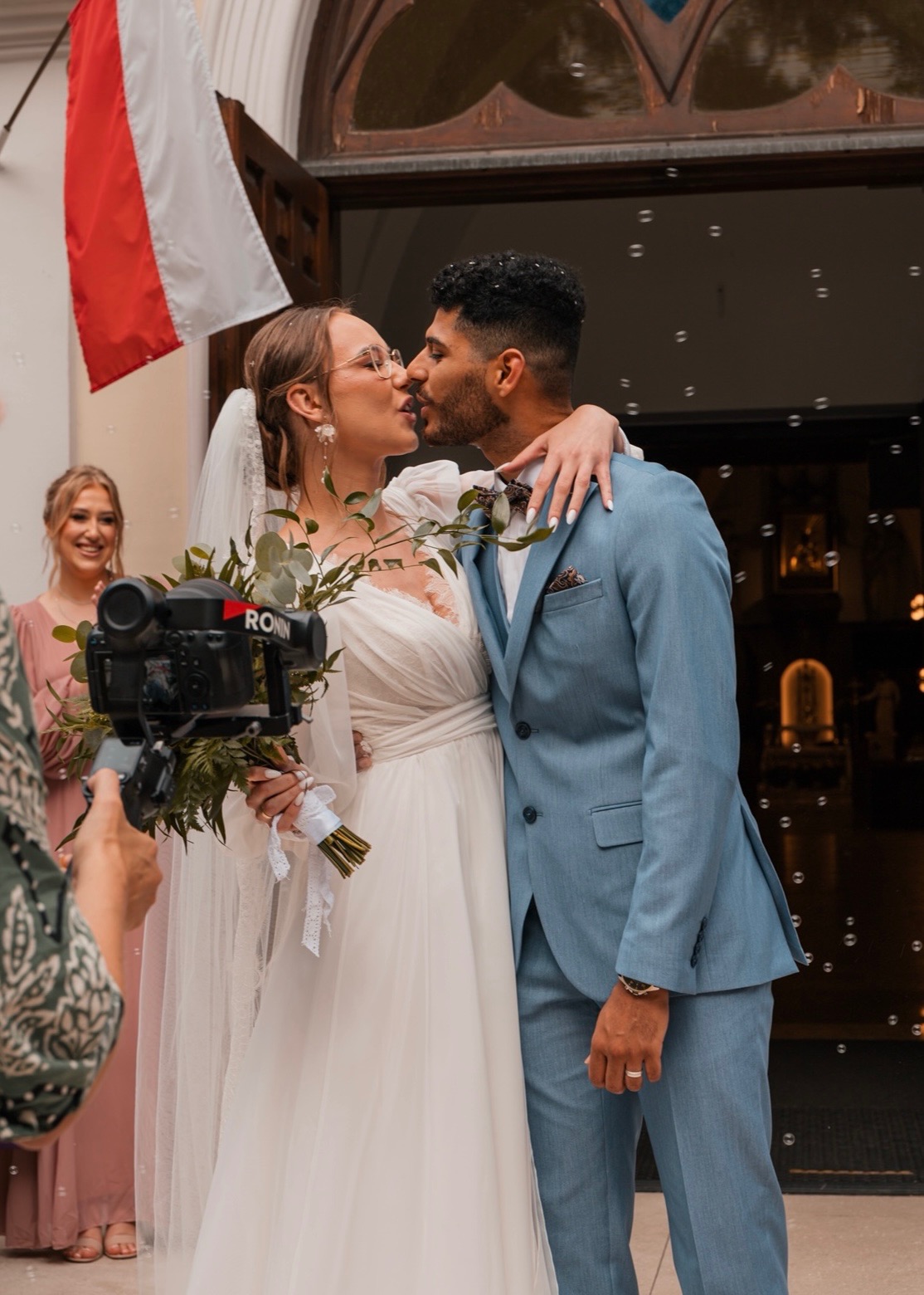 Pocałunek pary młodej na tle kościoła z flagą Polski i bańkami mydlanymi, filmowany z użyciem stabilizatora Ronin. Panna młoda w okularach i z bukietem.