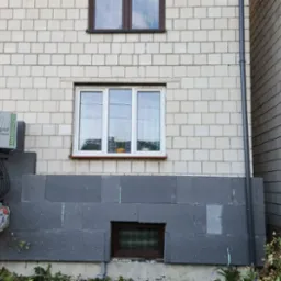 Fragment elewacji budynku z oknami, częściowo ocieplony grafitowym styropianem, na balkonie paczka styropianu Mega Styro