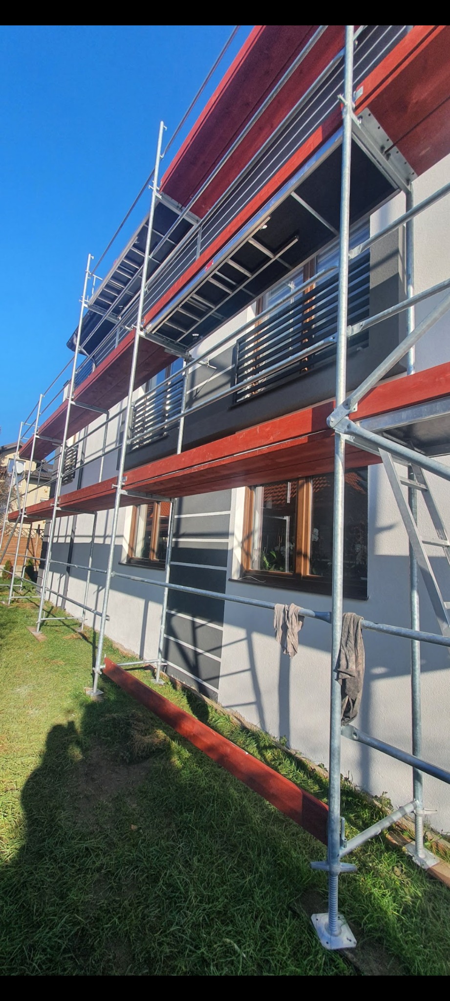 Dom jednorodzinny w trakcie prac elewacyjnych, widoczne rusztowania z czerwonymi deskami, fragment ogrodu i cień osoby robiącej zdjęcie.