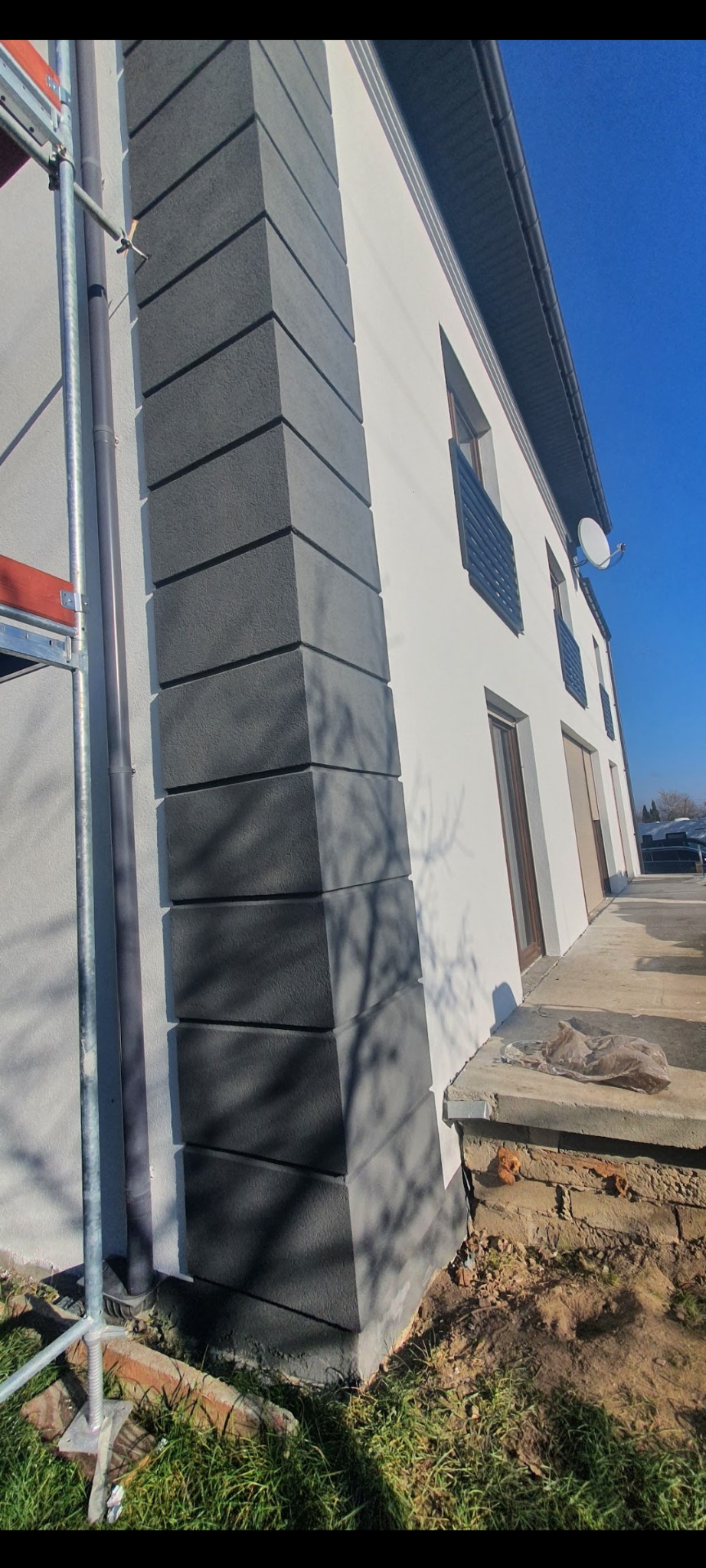 Wykończony narożnik budynku z szarą dekoracyjną elewacją w blokach, biała ściana, widoczne rusztowanie, rynna i fragment trawnika.