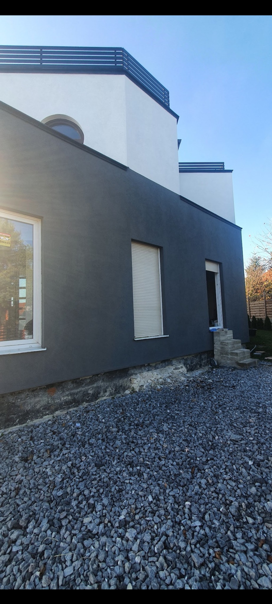 Nowoczesny dom po renowacji elewacji, z antracytową ścianą boczną i białym wykończeniem górnych kondygnacji, widoczne okno z roletą i wejście z kamiennymi schodami, teren wokół wysypany szarym żwirem.