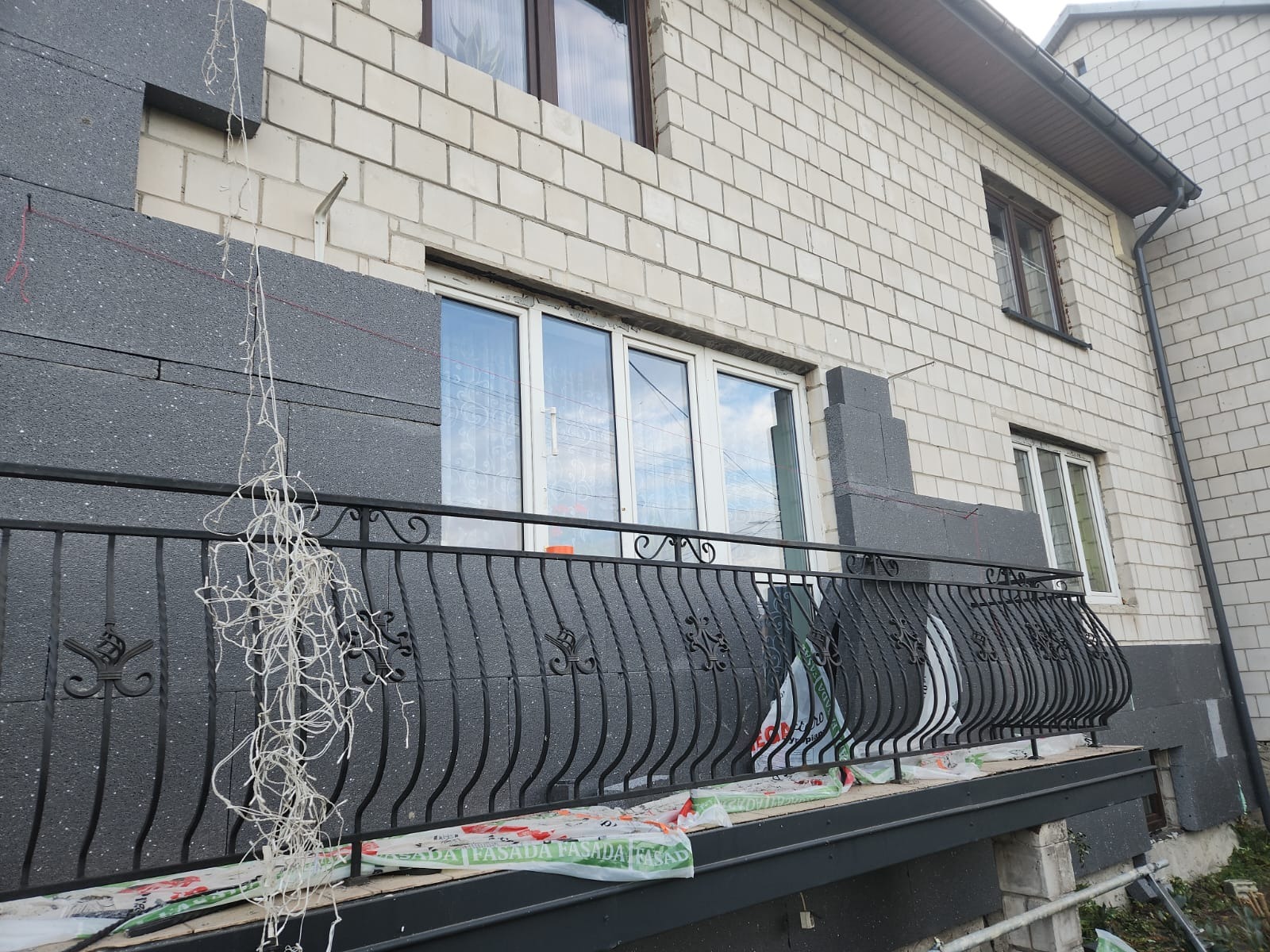 Budynek z białej cegły z częściowo ocieploną elewacją grafitowym styropianem, widoczny balkon z ozdobną, metalową balustradą i zwisającymi kablami oraz folie zabezpieczające.