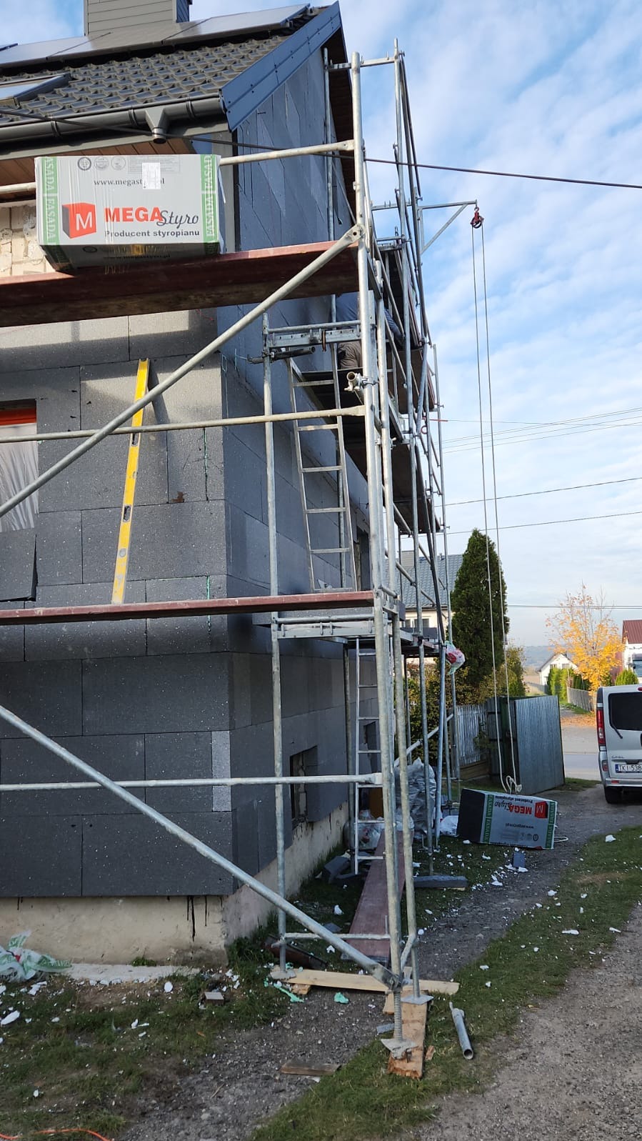 Dom w trakcie ocieplania szarą wełną mineralną, widoczne rusztowanie, paczka styropianu Mega Styro na rusztowaniu, poziomica przyłożona do ściany, resztki materiałów budowlanych na ziemi.