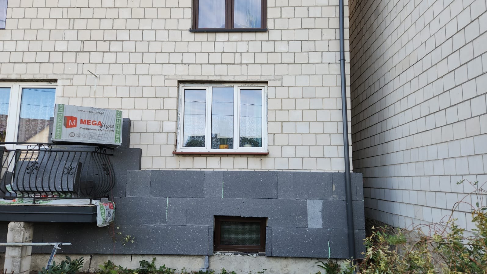 Fragment elewacji budynku z oknami, częściowo ocieplony grafitowym styropianem, na balkonie paczka styropianu Mega Styro
