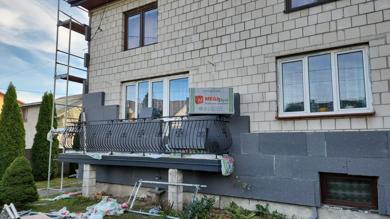 Dom w trakcie ocieplania szarym styropianem, widoczne rusztowanie, balkon z czarną balustradą i paczki styropianu Mega Styro.