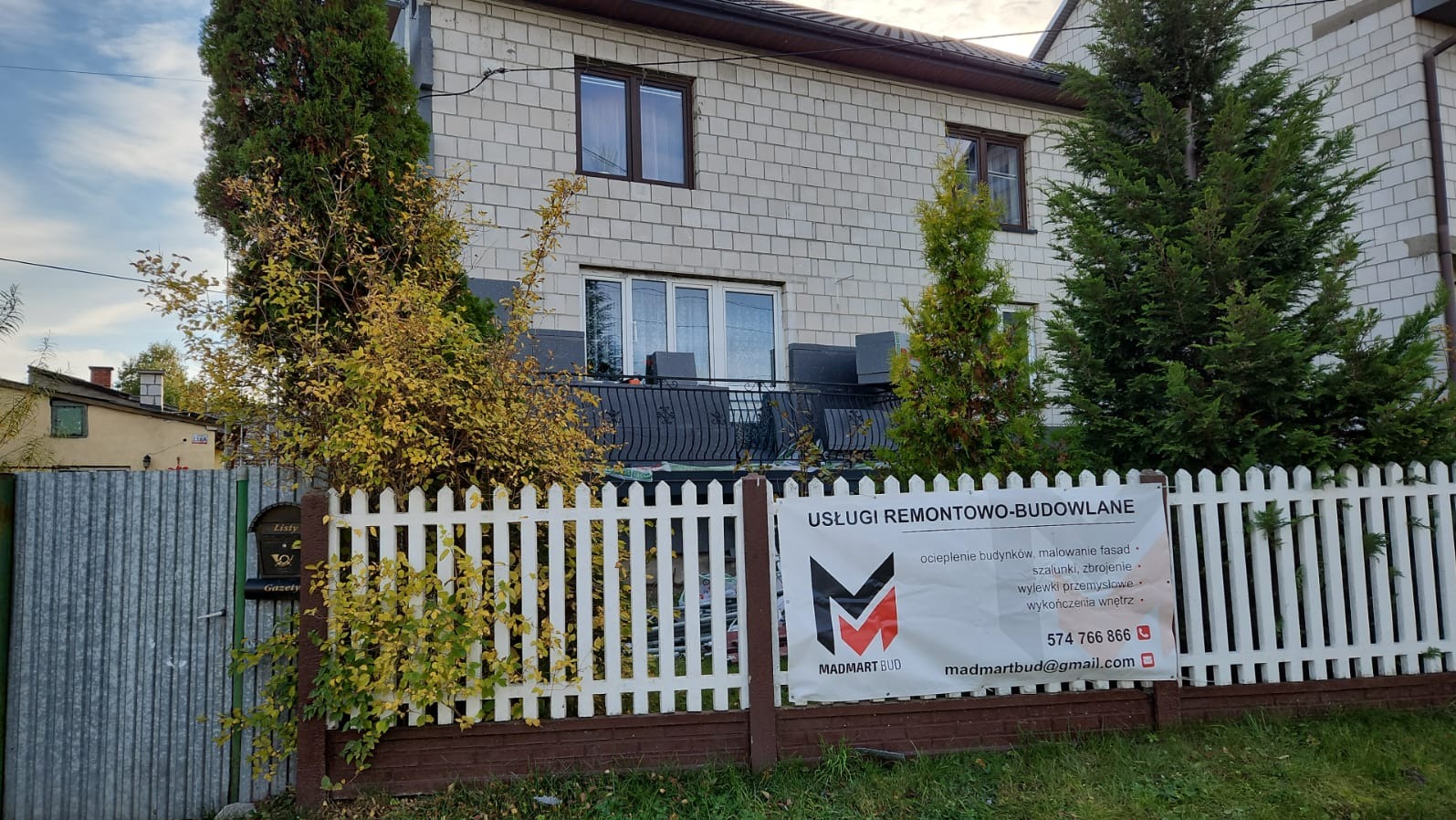 Dwupiętrowy dom z białej cegły z balkonem, widoczny zza białego płotu z reklamą usług remontowo-budowlanych Madmart Bud, otoczony drzewami i krzewami.