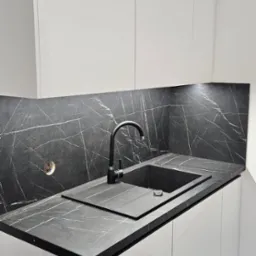 Minimalistyczna kuchnia: białe, gładkie szafki z czarnym blatem imitującym marmur i czarnym zlewem z baterią.