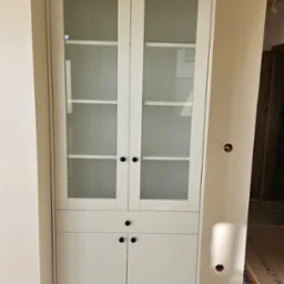 Biała przeszklona szafa z szufladami i szafkami w trakcie montażu, widoczne niedokończone wykończenie ścian i instalacja elektryczna.