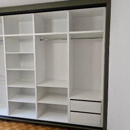 Bomax - Wnętrze białej, otwartej szafy wnękowej z przesuwnymi drzwiami, widoczne półki, drążki i szuflady, rama w kolorze khaki, parkiet na podłodze.