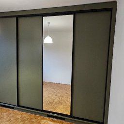 Bomax - Zielona szafa przesuwna z lustrem w pokoju z parkietem, widoczne gniazdko elektryczne i lampa sufitowa.