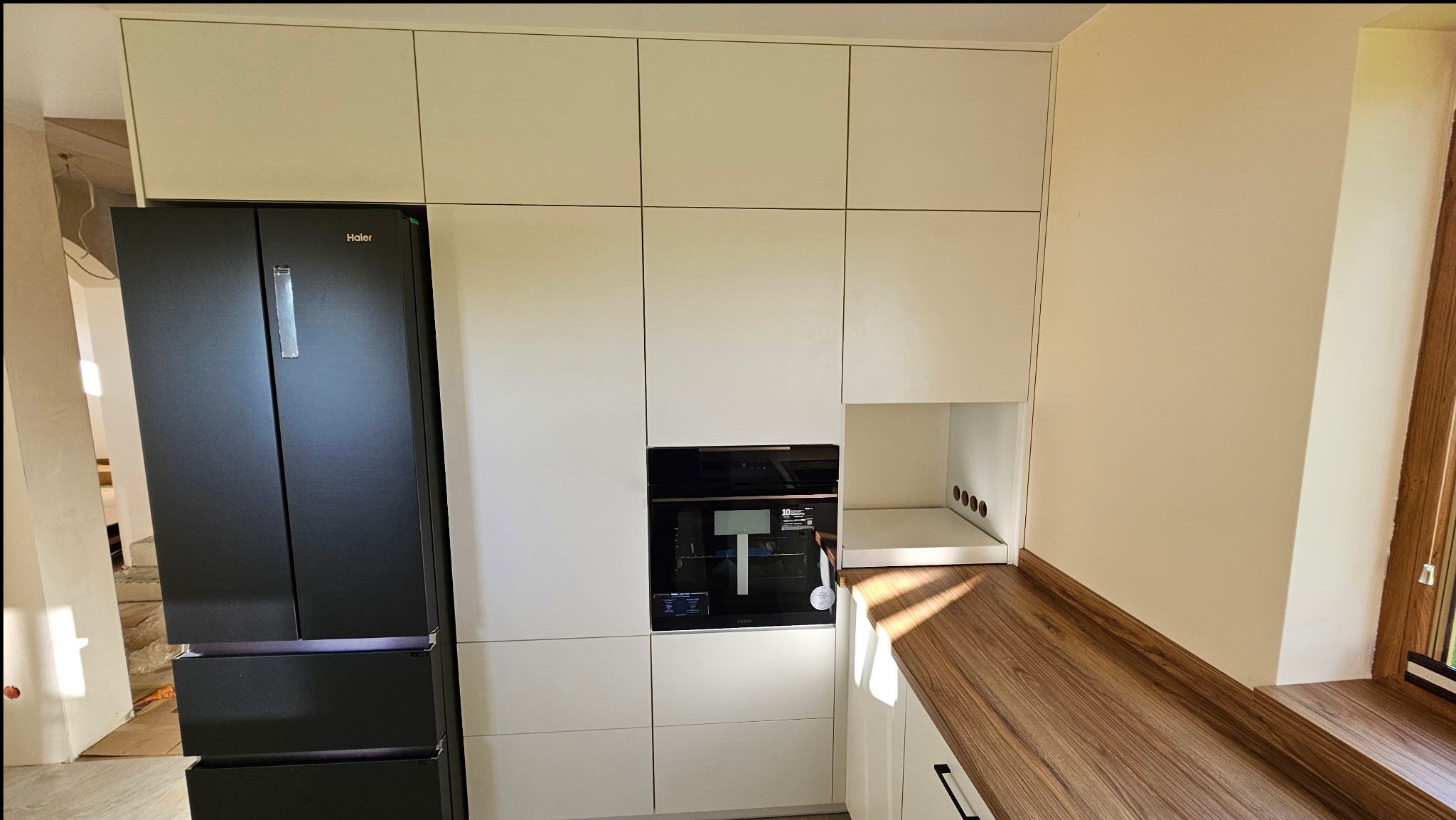 Nowoczesna zabudowa kuchenna z białymi frontami, czarnym lodówko-zamrażarką Haier i drewnianym blatem, z widocznym piekarnikiem w zabudowie i otwartą półką z otworami wentylacyjnymi.